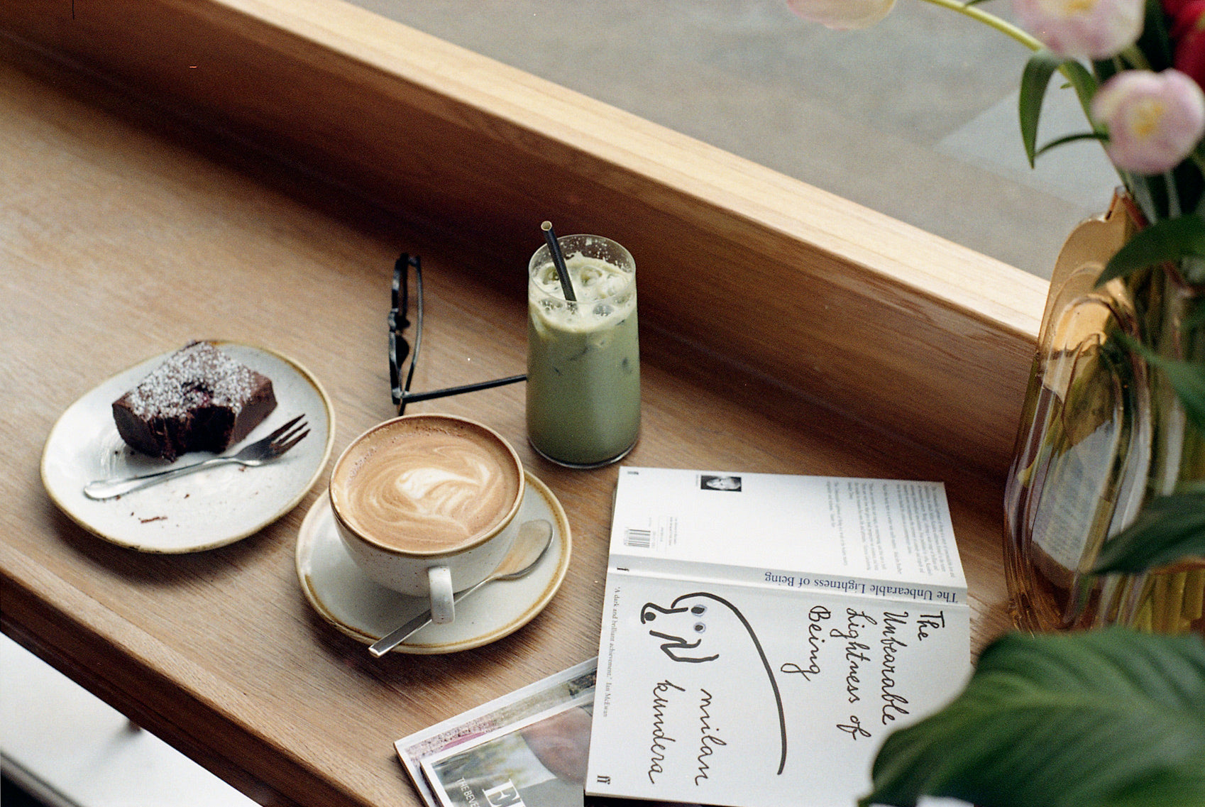 A Coffee cup with a latte, an iced matcha drink in a glass and a chocolate brownie with a part missing on a plate with a fork.  All on a wooden table with a book next to them